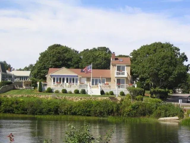 A tan waterfront house with a two-story section, an American flag, and surrounding trees sits above a calm pond.