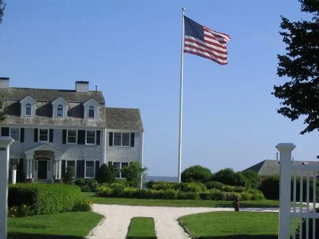 A large American flag flies on a tall pole in front of a white, multi-story coastal home with a gravel driveway.