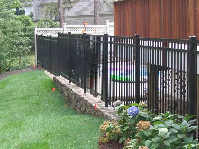 A black metal fence sits atop a stone retaining wall in a grassy backyard, bordering a pool area with a wooden wall.
