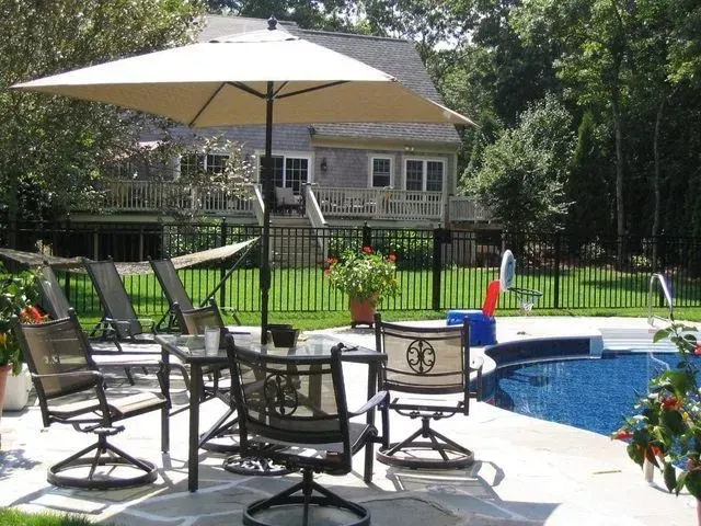 A patio with a table, chairs, and an umbrella, located next to a swimming pool in a grassy backyard with a house behind.