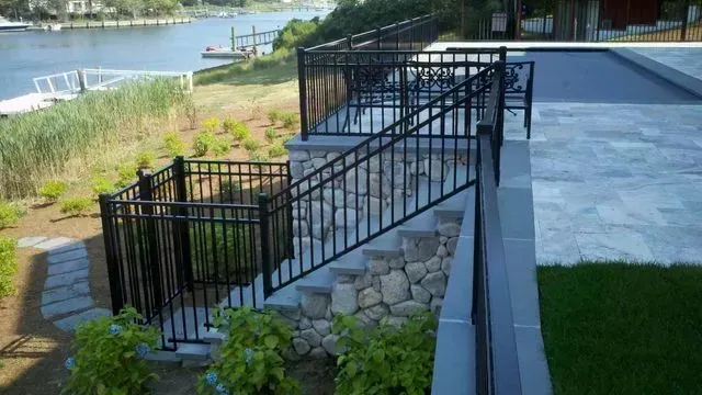 Black metal railing along stone steps leading from a paved patio down to a grassy area next to a waterway.