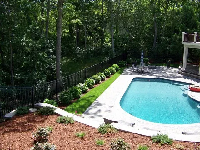 A backyard pool area with a stone patio, a green lawn, a row of shrubs along a black fence, and a wooded background.
