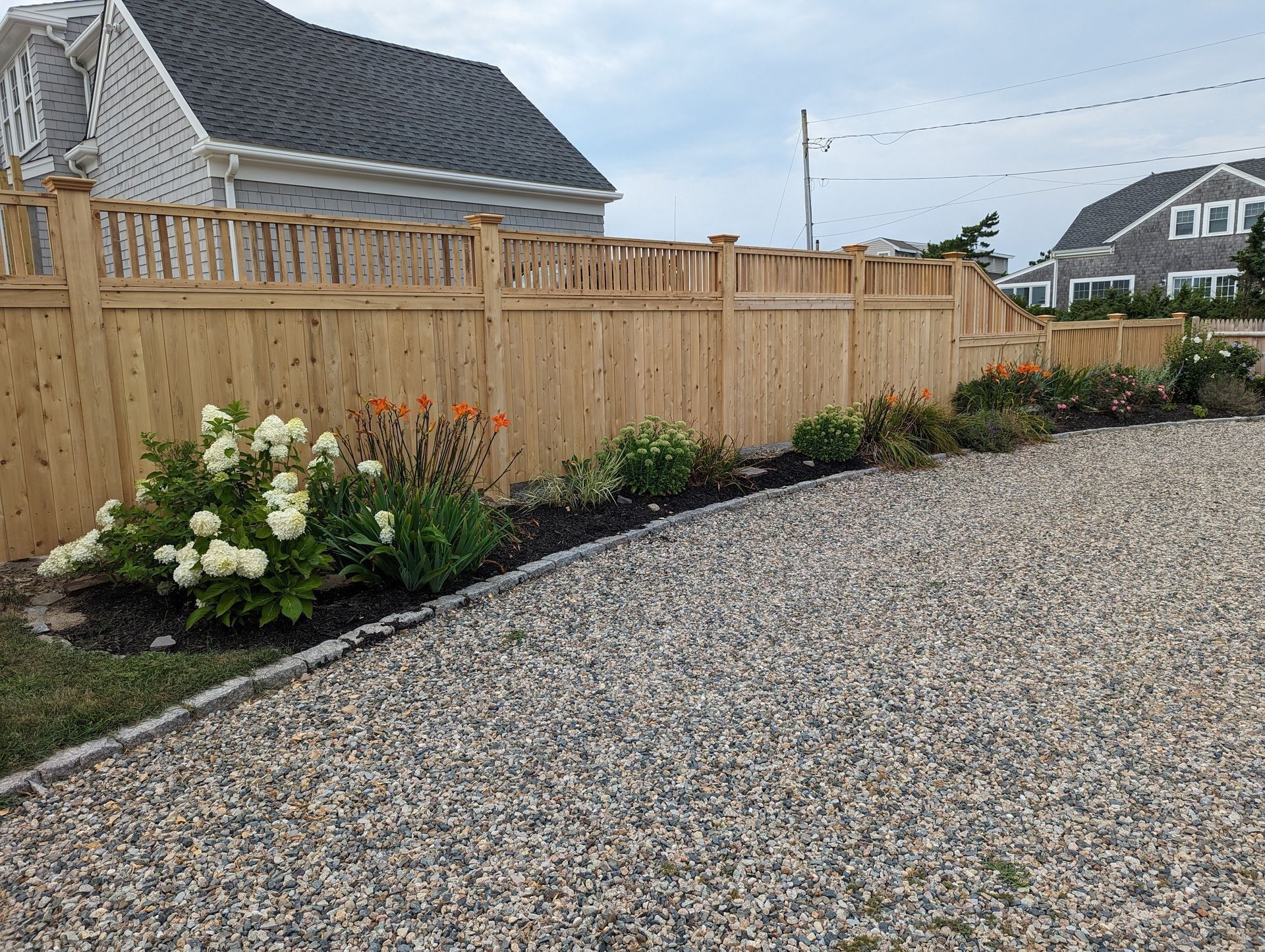 A wooden fence borders a gravel driveway, featuring a garden bed with white hydrangea bushes and orange flowers.