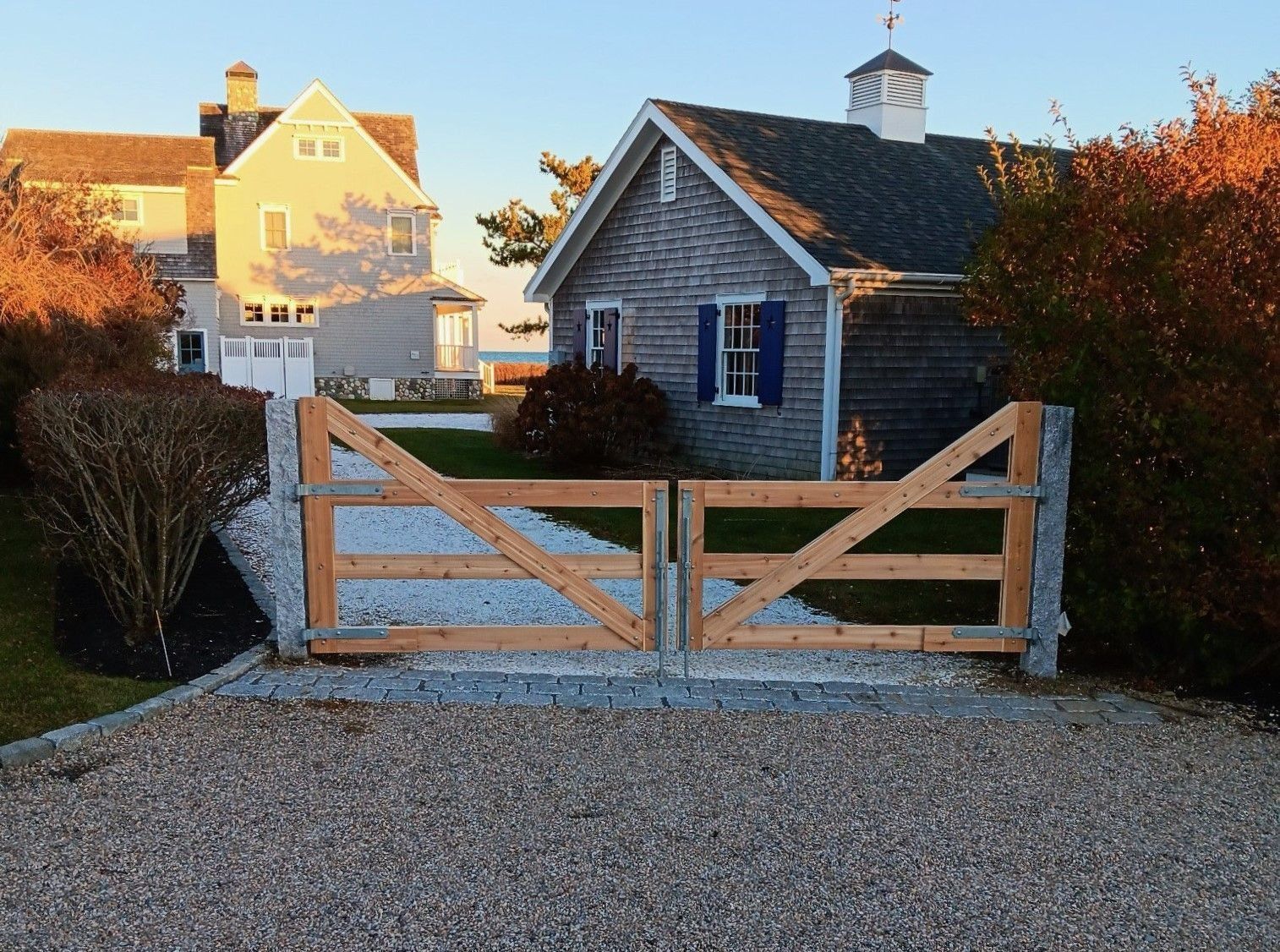 A wooden driveway gate stands before a shingled cottage and a large house at sunset, with light reflecting off the facade.