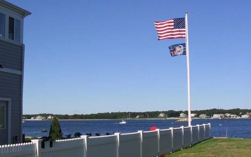 An American flag and a blue decorative flag wave on a tall pole by a white fence overlooking a bay on a sunny day.