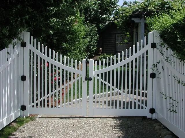 A white vinyl double gate with diagonal bracing, set in a matching picket fence on a gravel driveway.