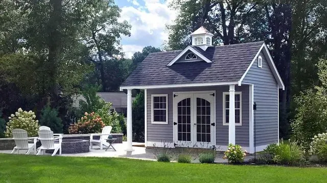A gray shed with a cupola and front porch, set in a backyard near a stone fire pit with white chairs.