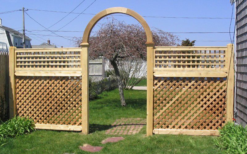 A wooden garden fence with lattice panels and a central arched entrance, set in a grassy backyard with a tree behind it.