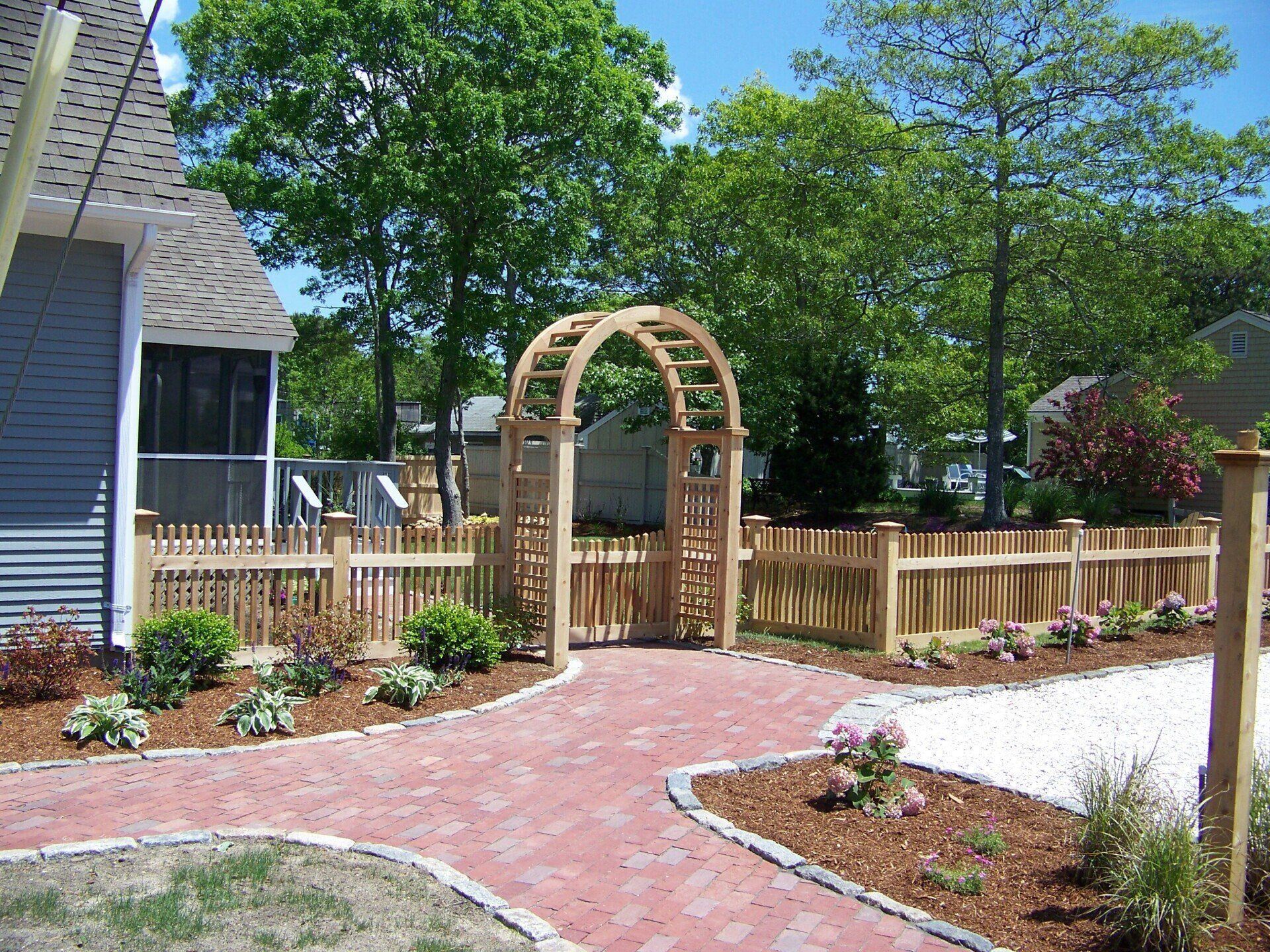 A red brick path leads to a wooden garden arch and picket fence in a sunny residential yard with green trees.