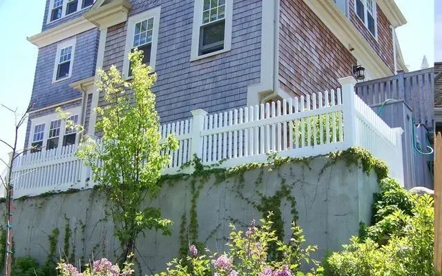 A white picket fence atop a gray stone retaining wall in front of a blue shingled house on a sunny day.
