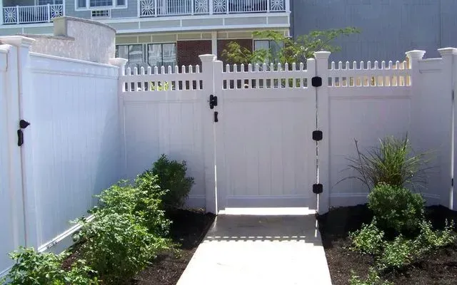 A white vinyl fence with a matching gate set against a building, with garden plants in the foreground.