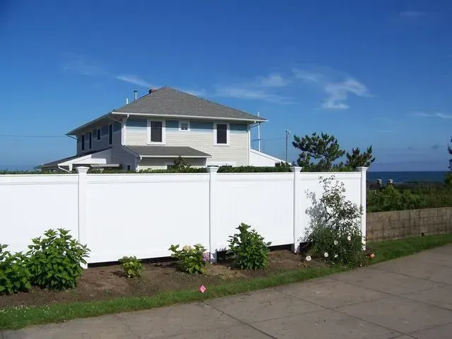 A house with light-colored siding and a gray roof stands behind a tall, white privacy fence on a sunny day.