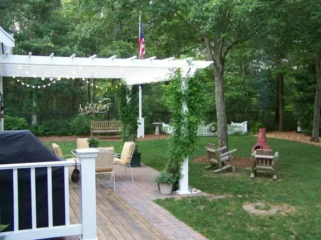 A backyard patio featuring a white pergola, wooden deck, outdoor seating, and a fire pit, surrounded by trees and greenery.