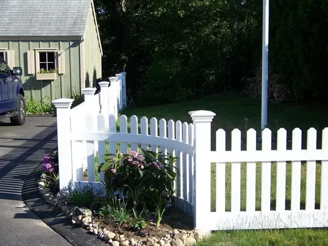 A curved white vinyl picket fence stands in a grassy yard next to a driveway and a shed, with small plants in the foreground.