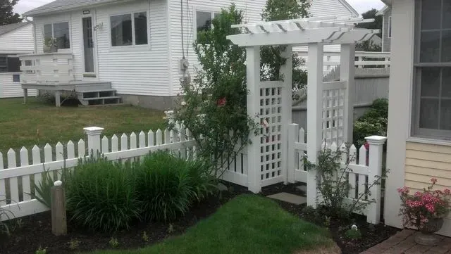 A white picket fence and trellis gate frame a garden bed with green plants in front of a white house.