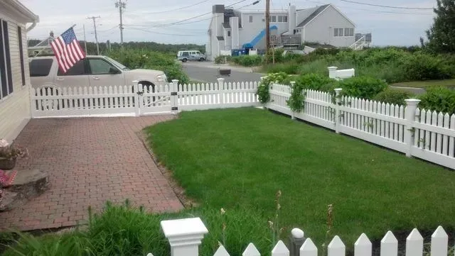 A small grassy yard bordered by a white picket fence, with a brick patio, a parked car, and a house in the background.