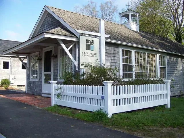 A gray, shingled small building with a white picket fence, a bell tower, and a sign in a grassy setting.