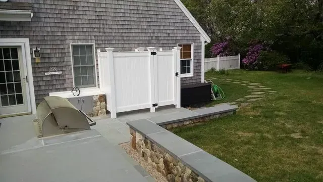 A gray-shingled house with a white privacy fence, stone retaining wall, patio, and a backyard with a green lawn.