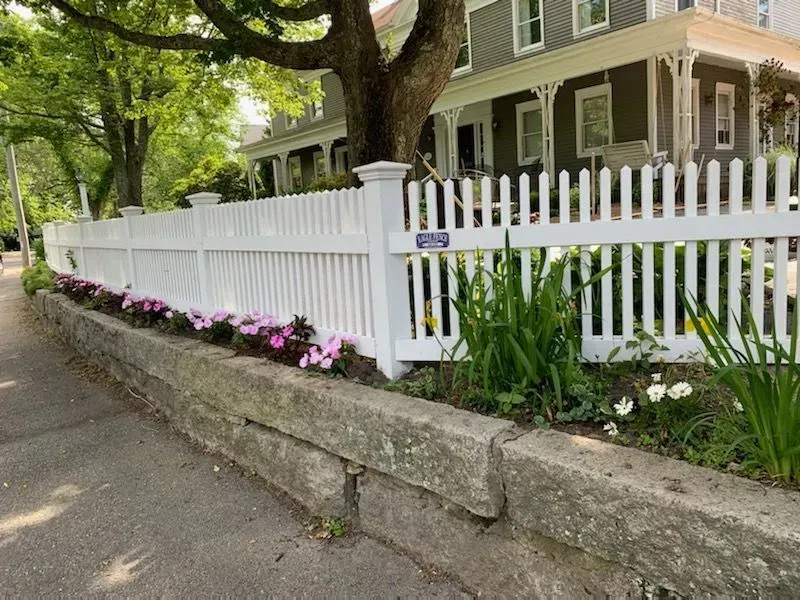 A white picket fence runs along a stone retaining wall in front of a house, with pink flowers planted in the garden bed.