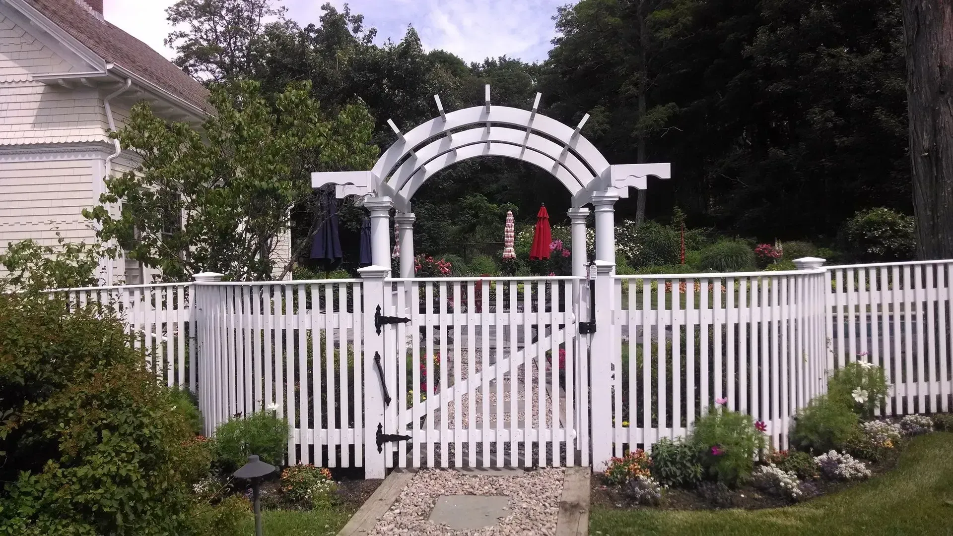 A white picket fence and a gated arched arbor lead to a landscaped garden beside a house.