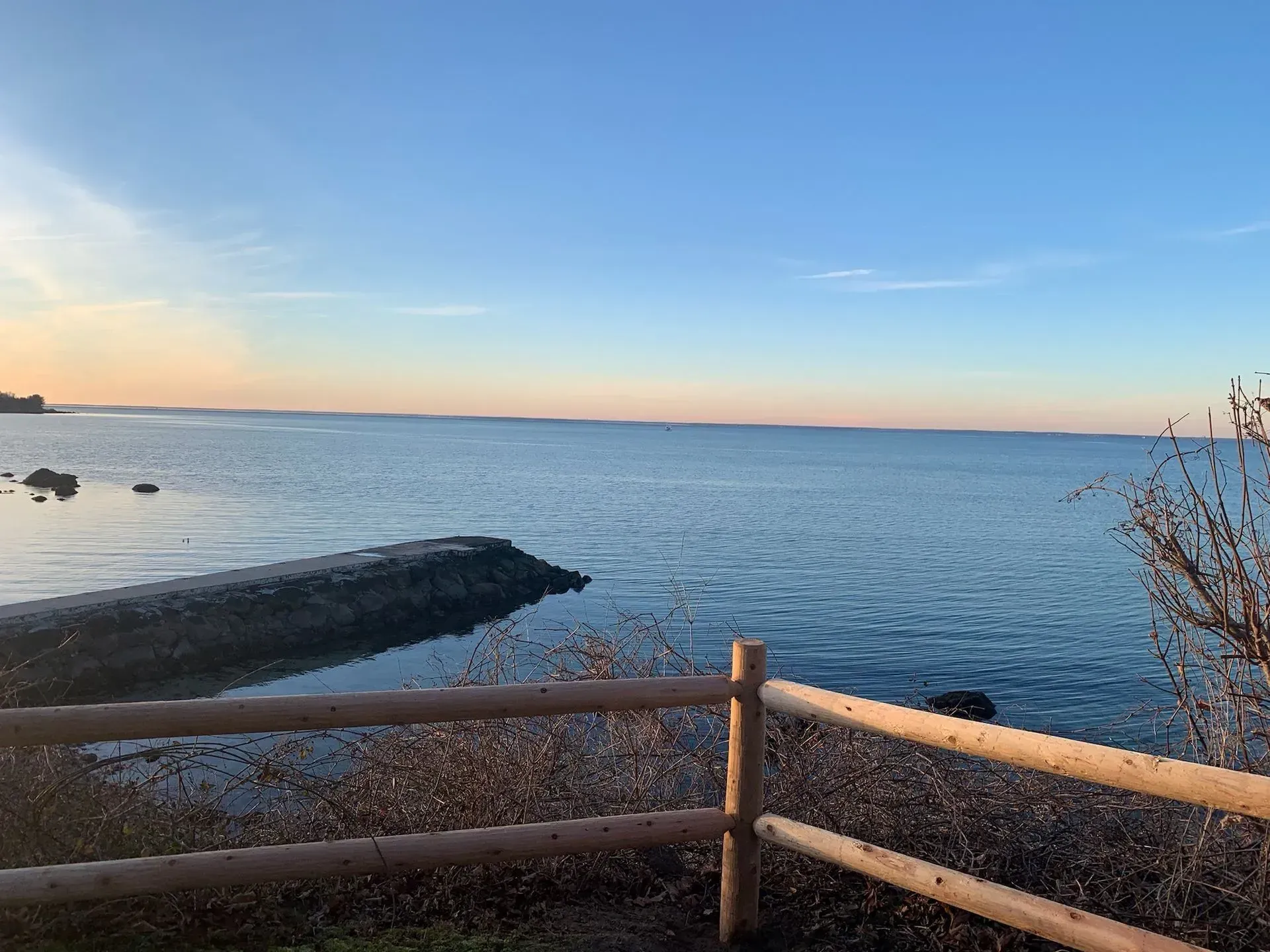 A wooden fence overlooking a calm bay at sunset with a stone jetty stretching into the water.