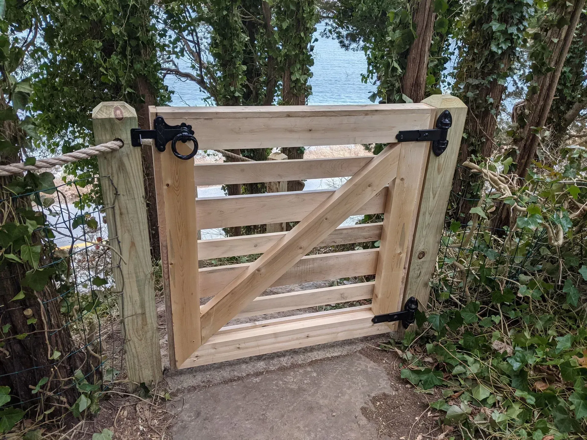 A wooden gate with black hinges and a latch stands on a path, surrounded by trees with a coastal view in the background.