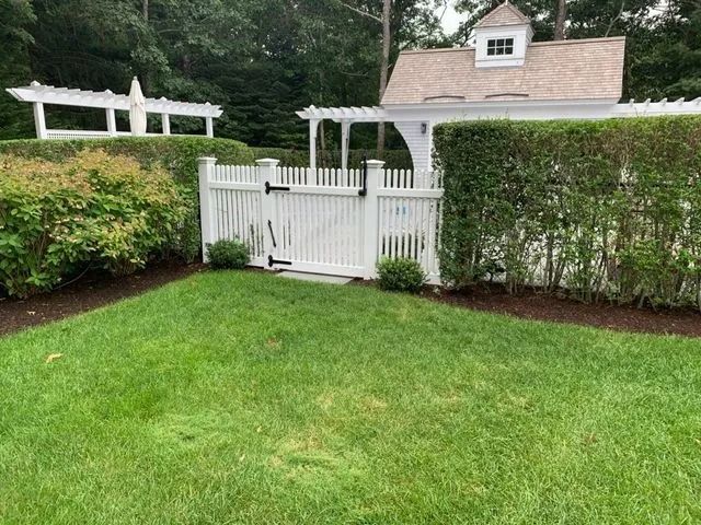 A white picket gate and fence line a lush green lawn, leading to a white building with a cupola and a wooden trellis.