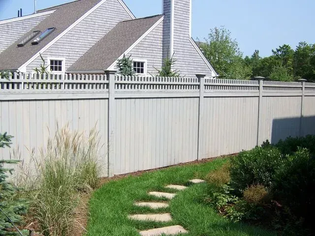 A tall, light-colored privacy fence with a decorative lattice top follows a stone pathway through a grassy backyard.