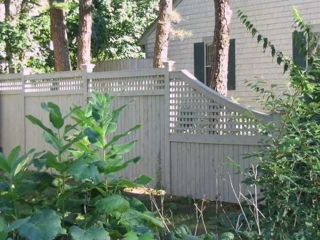 A grey privacy fence with lattice topping slopes downward in a garden setting next to a house.