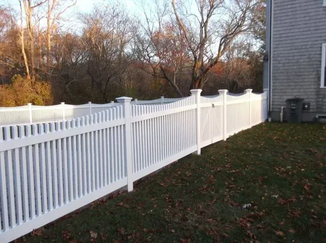 A white vinyl scalloped picket fence runs along the edge of a lawn next to a grey house with trees in the background.
