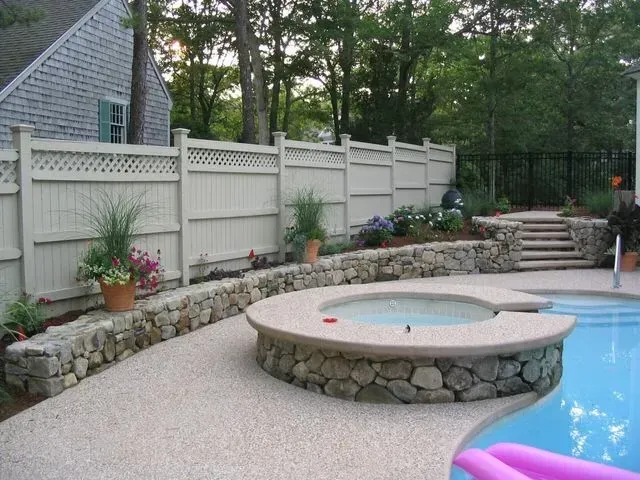 A swimming pool area with a circular spa, stone retaining walls, light tan decking, and a white privacy fence.