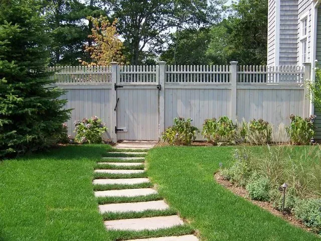 A stone stepping-stone path leads through a green lawn to a gate in a tall, light-colored vinyl fence.