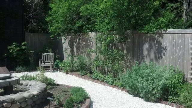 A wooden chair sits on a white gravel path in a backyard garden beside a stone fire pit and a tall wooden fence.