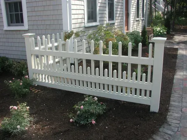 A white vinyl picket fence stands in a garden bed in front of a gray shingled house.