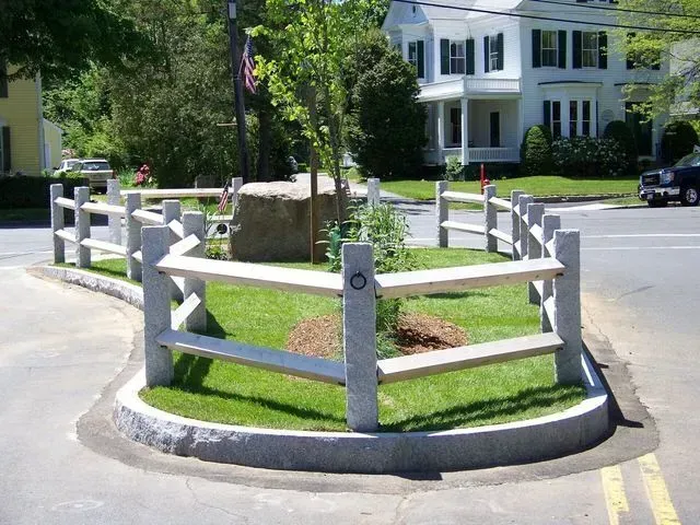A small, triangular traffic island landscaped with grass and a tree, enclosed by a granite post-and-rail fence.