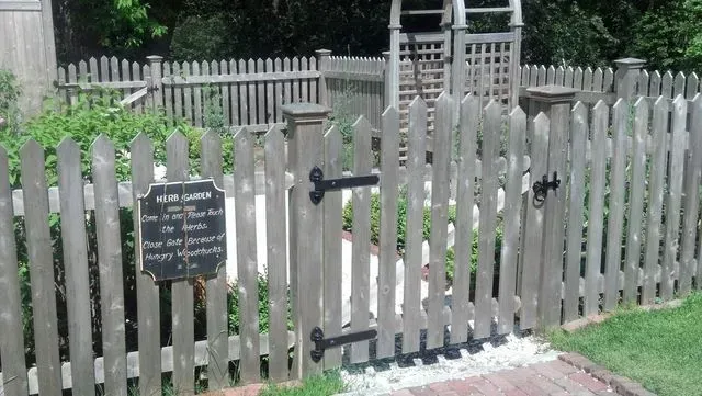A wooden picket fence and gated entrance to a garden, with a small sign hanging on the left panel.