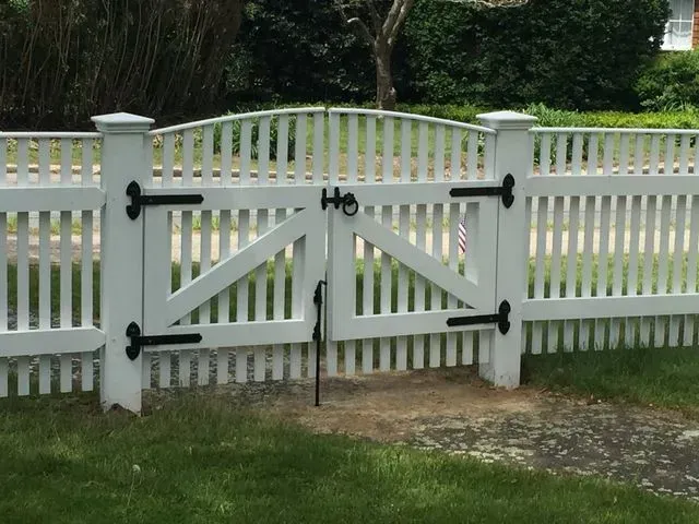 A white wooden picket fence with a double-gate entrance, featuring black metal hinges and a central gate latch.