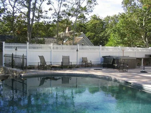 A backyard swimming pool area with patio chairs, a hot tub, and a white privacy fence against a wooded backdrop.