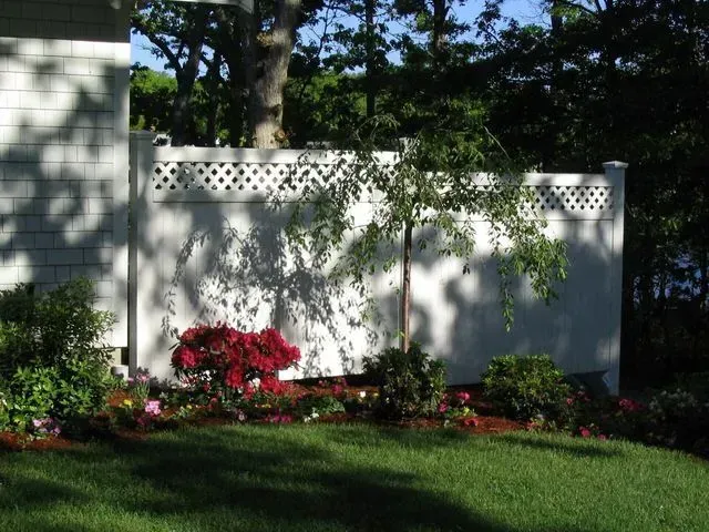 A white privacy fence with lattice top, fronted by a garden bed with red flowers, a small tree, and a green lawn.