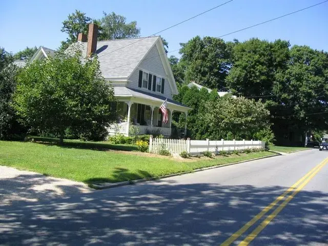 A light-gray, two-story house with a front porch, a white picket fence, and lush green trees along a paved country road.