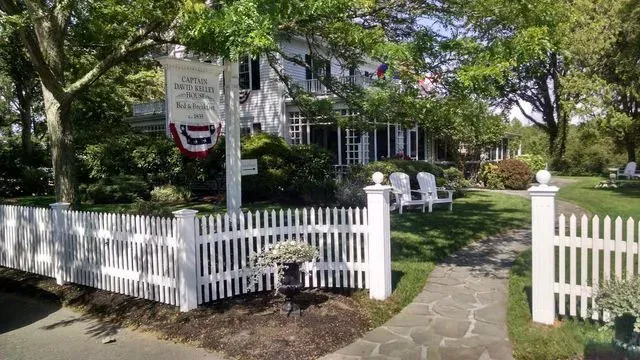 A white house with a picket fence and a wooden sign hanging from a post, set amidst lush green trees and a stone path.