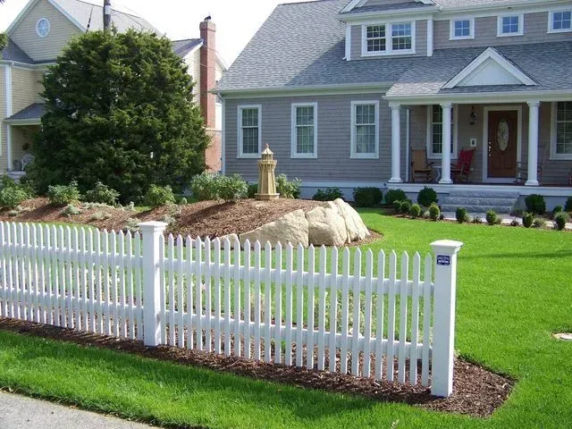 A white picket fence runs along a manicured lawn in front of a gray house with a decorative rock garden and a lighthouse.