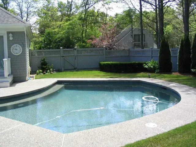 A backyard with a light-blue, custom-shaped swimming pool surrounded by a concrete deck and a gray lattice fence.
