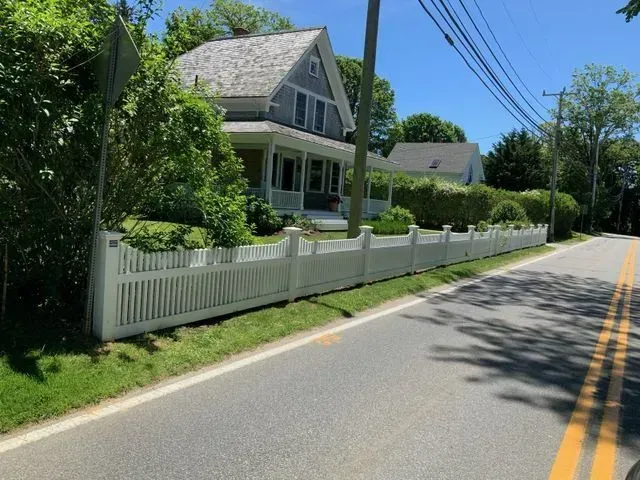 A gray house with a white picket fence sits behind a grassy lawn along a road on a sunny day.