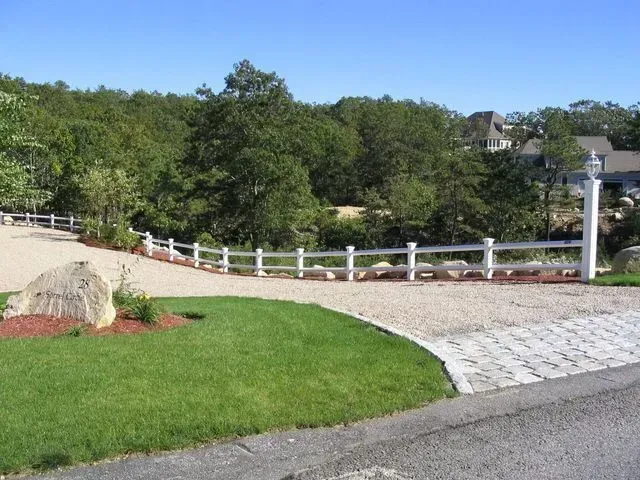 A white rail fence curves along a gravel driveway in front of a green, tree-filled landscape under a clear blue sky.