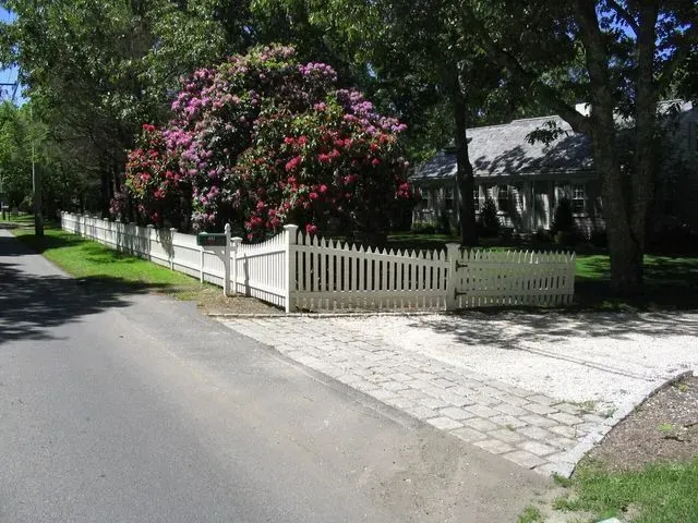 A white picket fence lines a driveway next to a road, with a large, flowering bush behind the fence in a residential area.