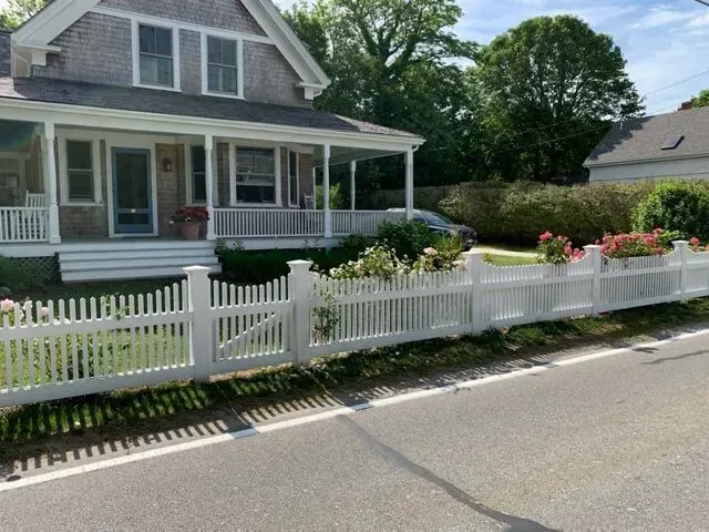 A white picket fence runs along a paved road in front of a gray-shingled house with a covered porch and flower boxes.