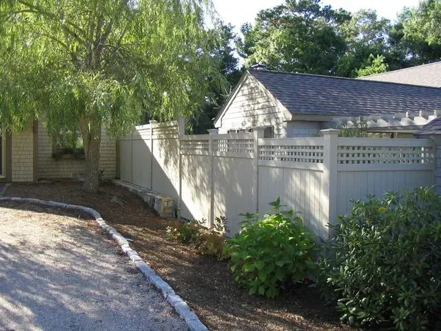 A tall tan privacy fence with a lattice top borders a driveway and landscaped yard next to a house.