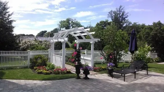 A white wooden arbor stands over a stone path in a sunny garden with a white picket fence, flower beds, and a patio bench.