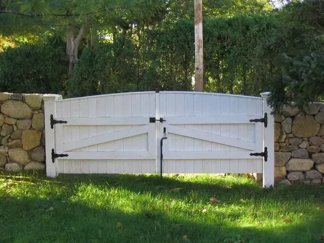 A white, double-gate with arched tops and black hinges, set within a stone wall against a backdrop of trees.